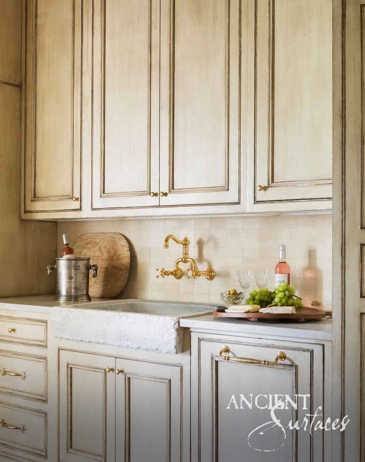 Reclaimed limestone farmhouse sink by Ancient Surfaces set into a French country kitchen with an unlacquered brass bridge faucet