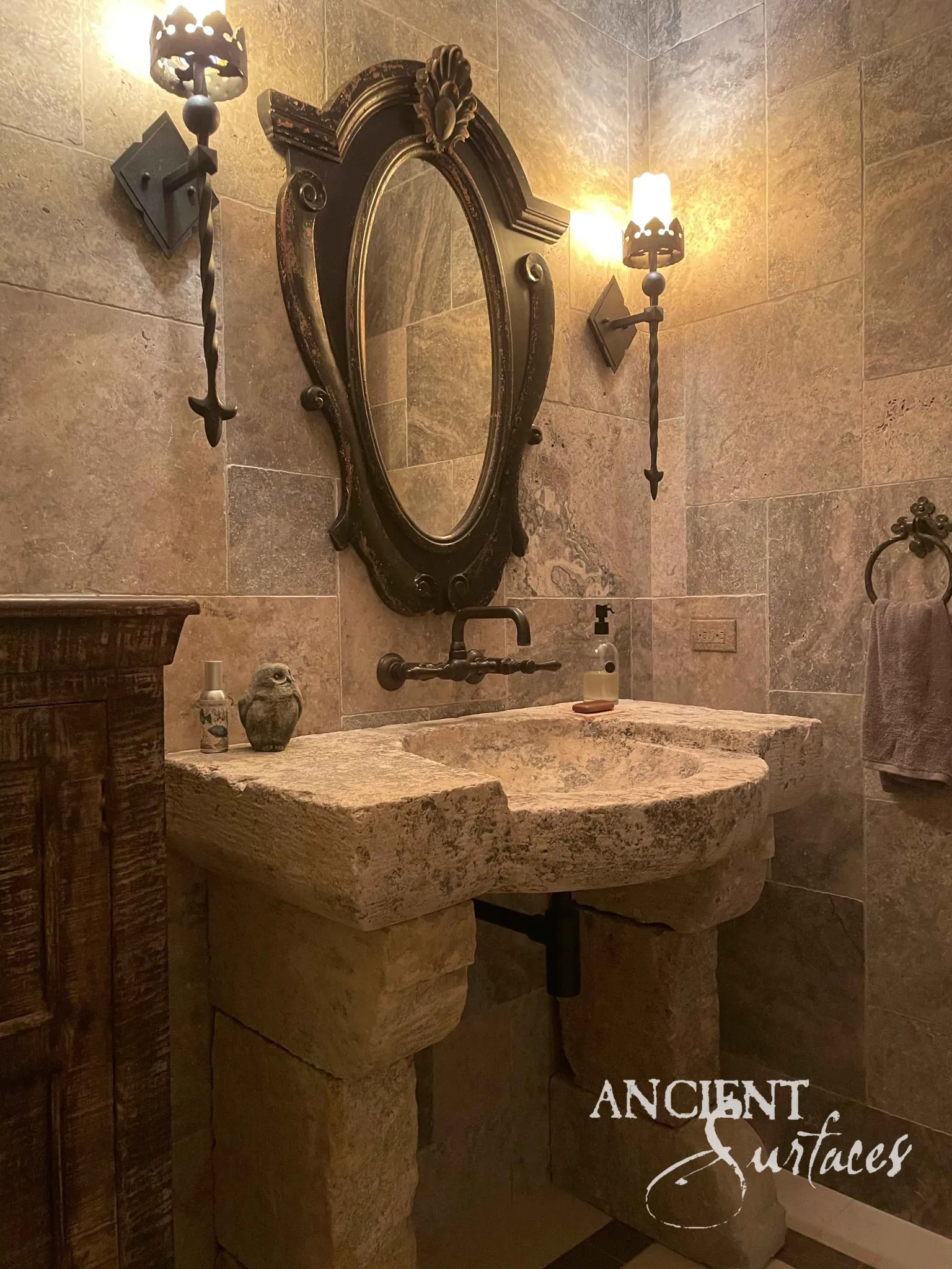 Antique limestone sink by Ancient Surfaces set on carved stone pedestals in a travertine powder room with wrought iron sconces